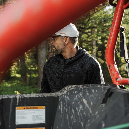 Man in a white cap wearing the Black Camo Blaze Tech Quarter Zip next to a muddy red utility vehicle in the woods