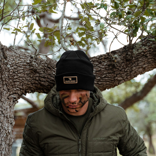 Man with face paint wearing the Black BURLEBO Beanie and an olive jacket under an oak tree