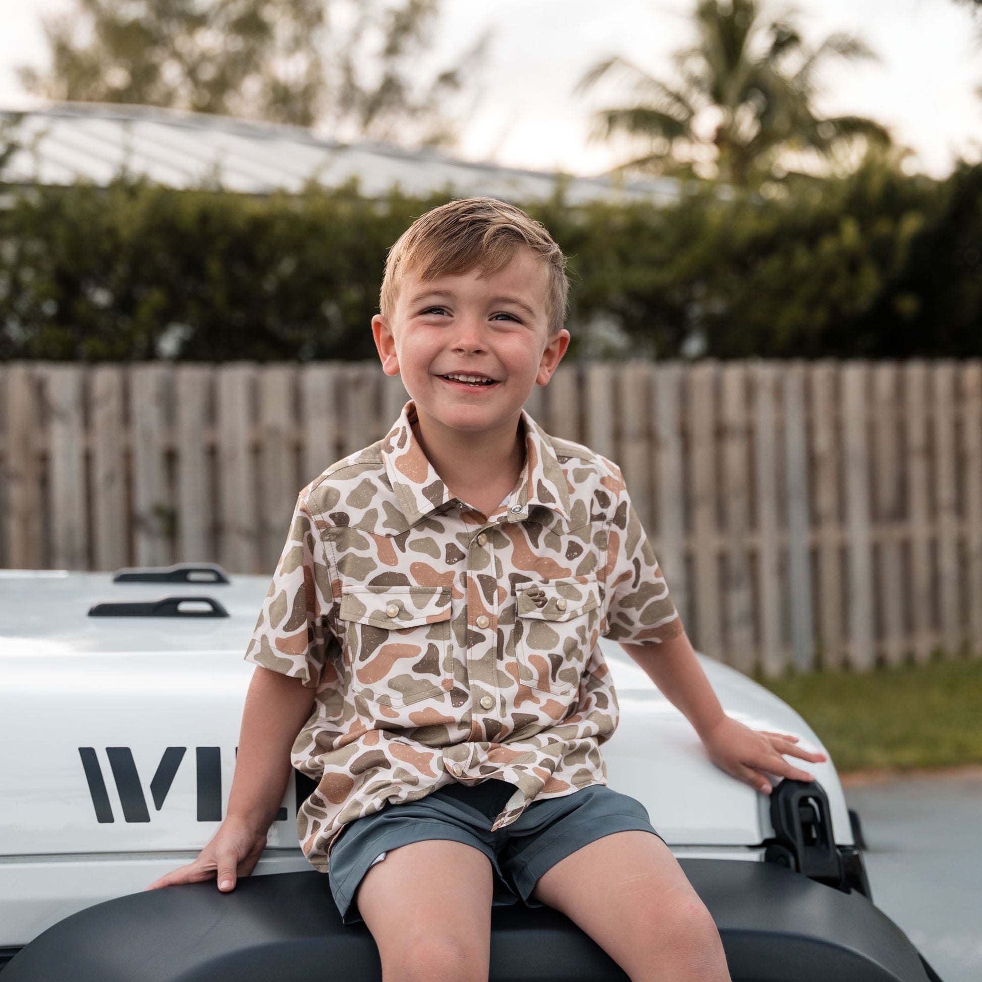 Child sitting on a vehicle roof with a camouflage shirt and shorts, outdoors.