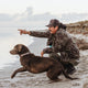 Man in camouflage jacket and cap with a brown dog by a body of water