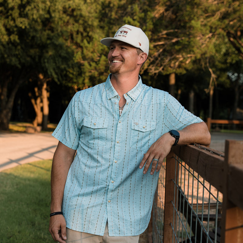 Man wearing a light blue patterned shirt and white cap standing outdoors with trees in the background