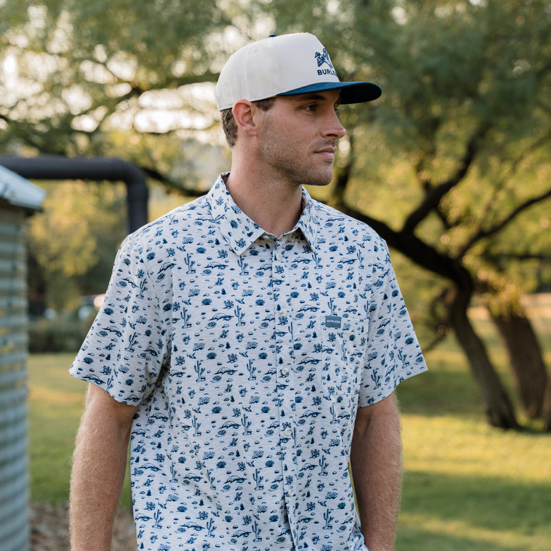 Man wearing a patterned shirt and cap outdoors with trees in the background