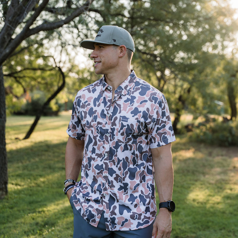 Man wearing a patterned shirt and cap standing in a park-like setting with trees and grass.