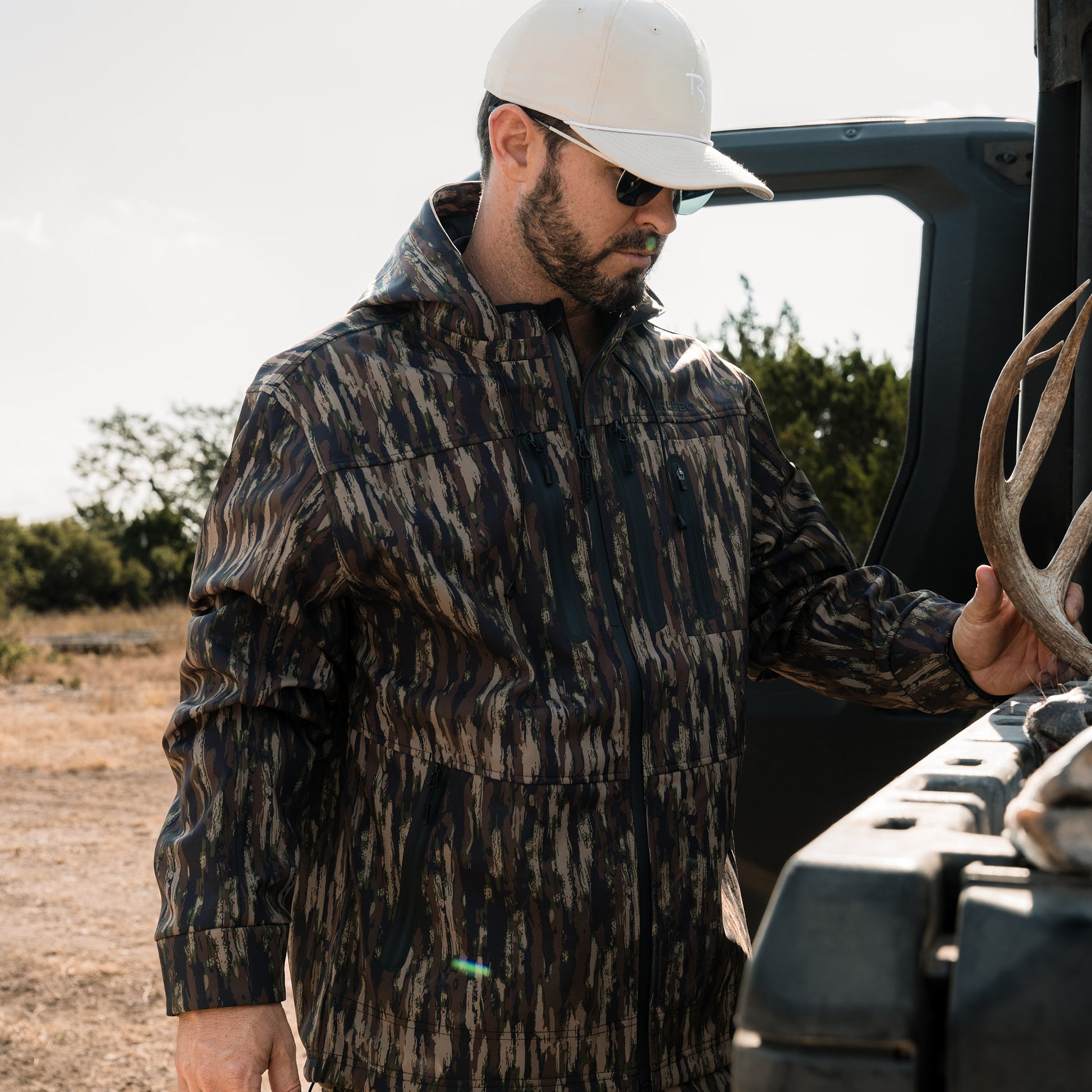 Man in camouflage jacket holding deer antlers next to an open truck door outdoors.