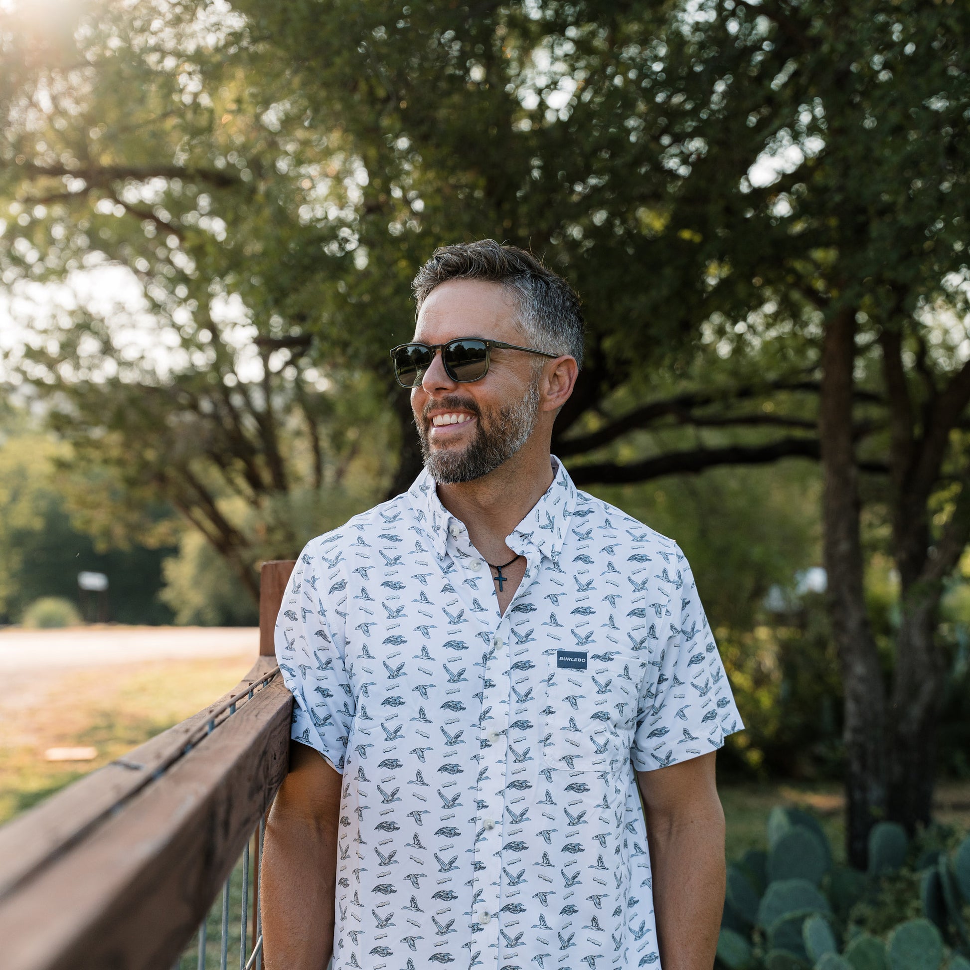 Man wearing sunglasses and a patterned shirt standing outdoors with trees and cacti in the background