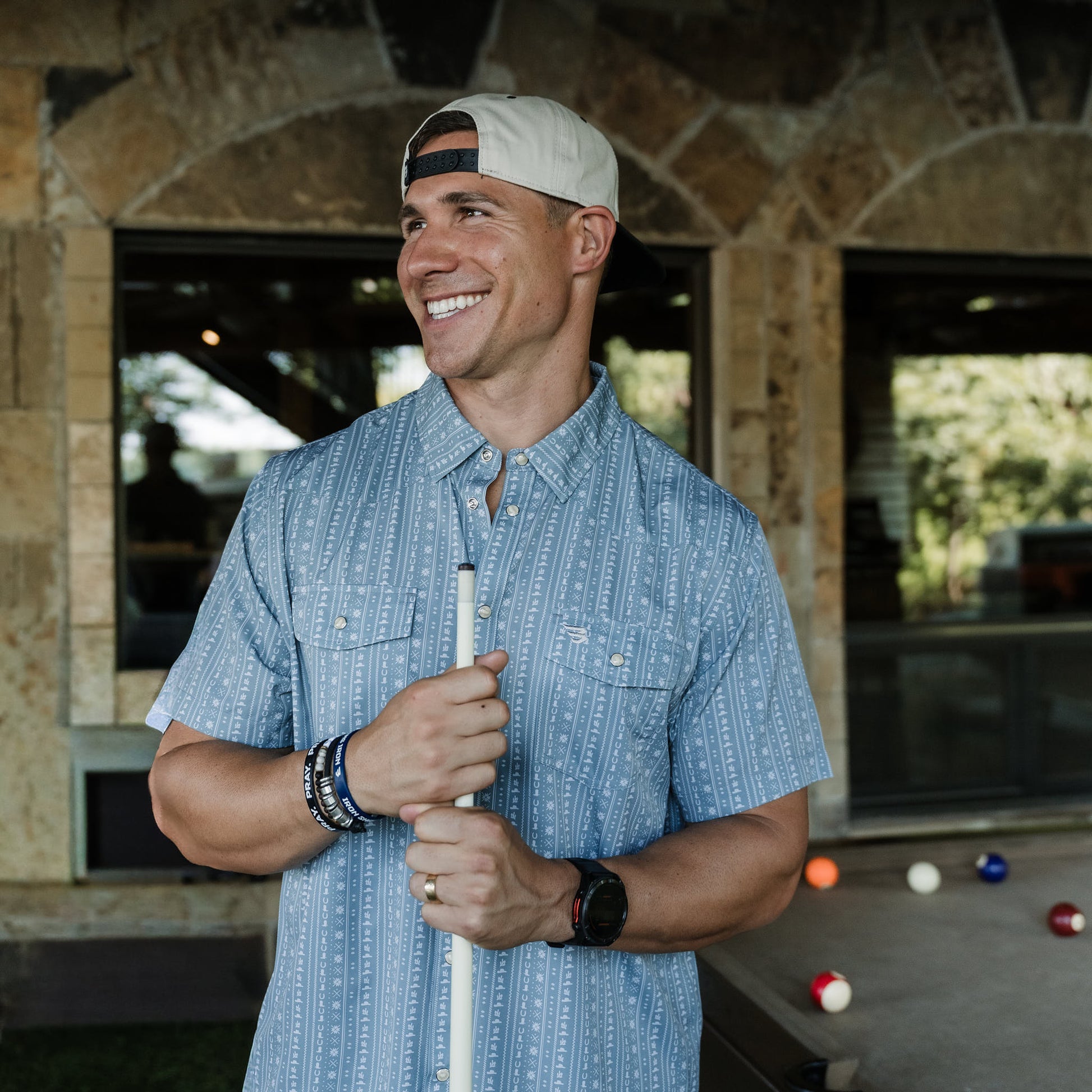 Man holding a pool cue outdoors with a stone building and pool table in the background