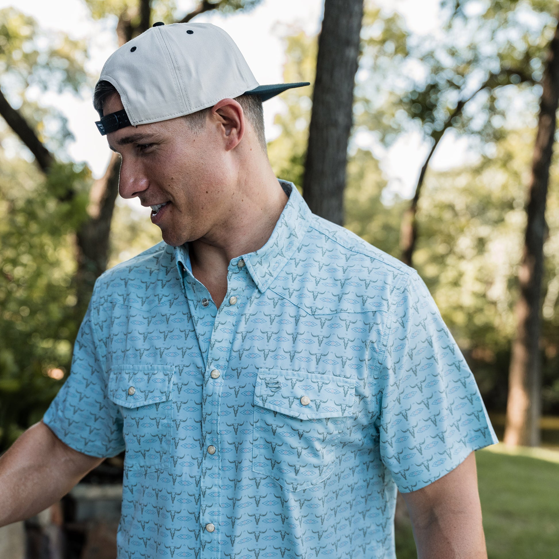 Man wearing a light blue patterned shirt and white cap outdoors with trees in the background