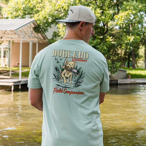 Man wearing a light green t-shirt with 'Burliebo Outdoors' logo and text, standing by a lake.
