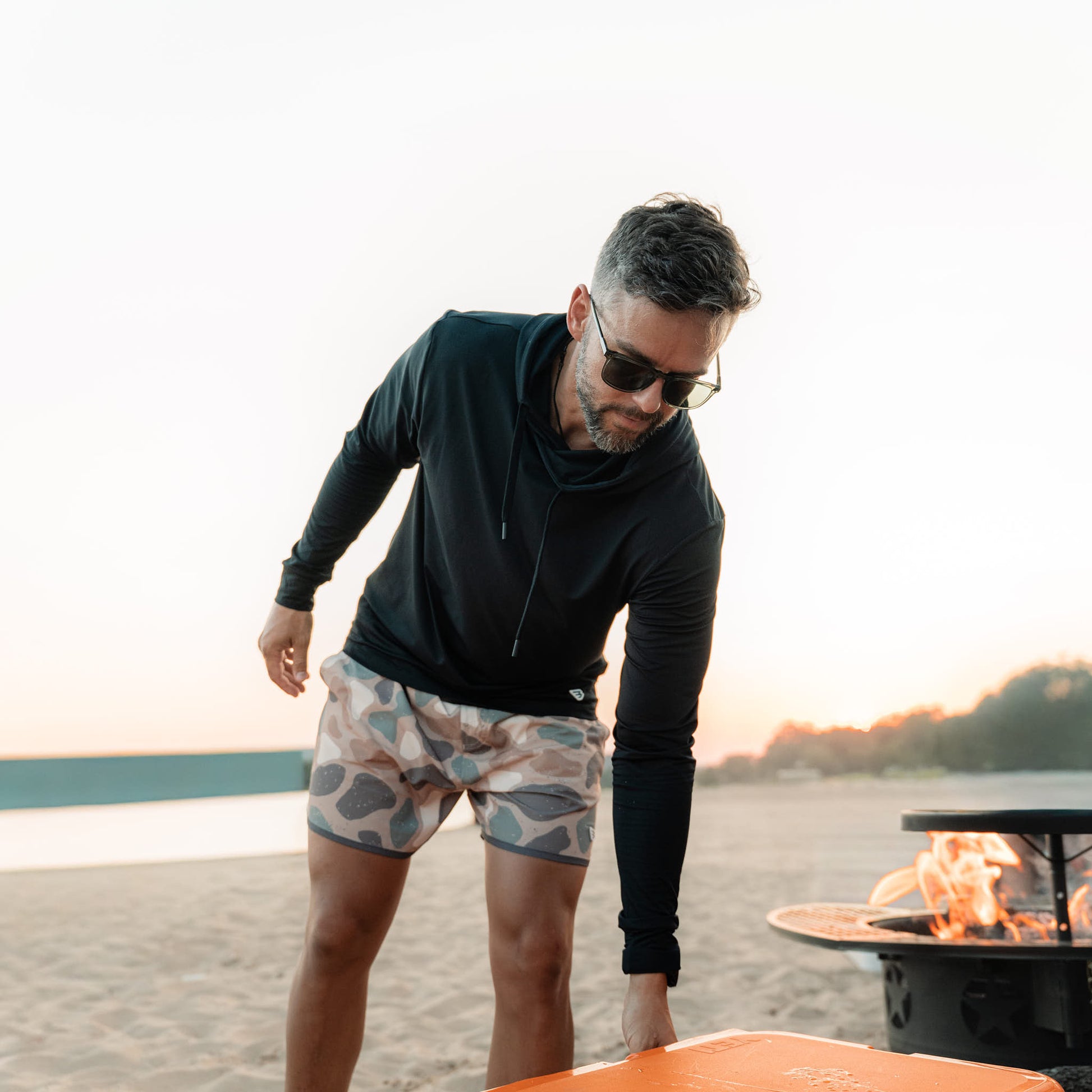 Man in black hoodie and patterned shorts by a fire pit at sunset