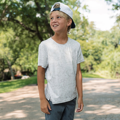 Young boy wearing a white t-shirt and cap outdoors with trees in the background