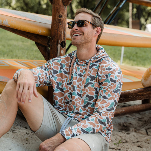 Man wearing a patterned hoodie sitting next to surfboards on a beach.