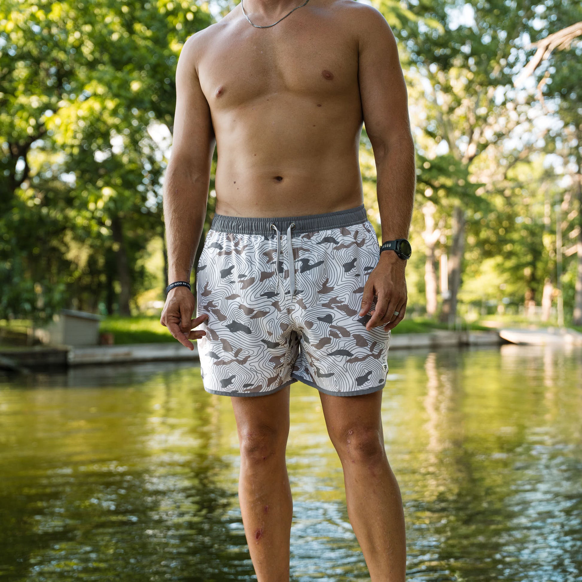 Man wearing patterned swim shorts standing by a body of water with trees in the background