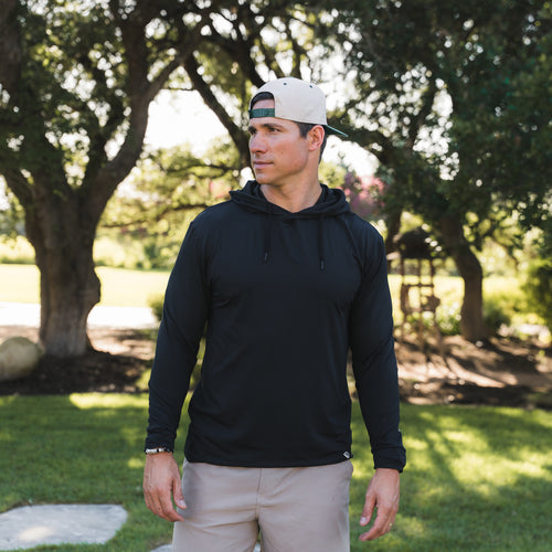 Man wearing a black hoodie and beige pants standing in a park-like setting with trees and grass.