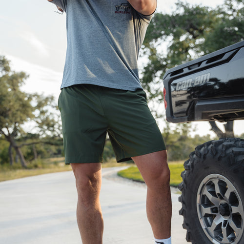 Person wearing green shorts and a gray shirt standing next to a black truck on a road with trees in the background.