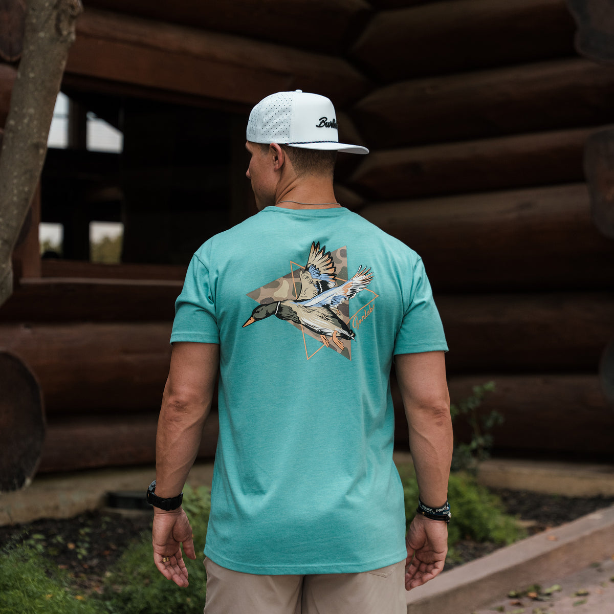 Man wearing a teal t-shirt with a graphic design, standing in front of stacked logs.