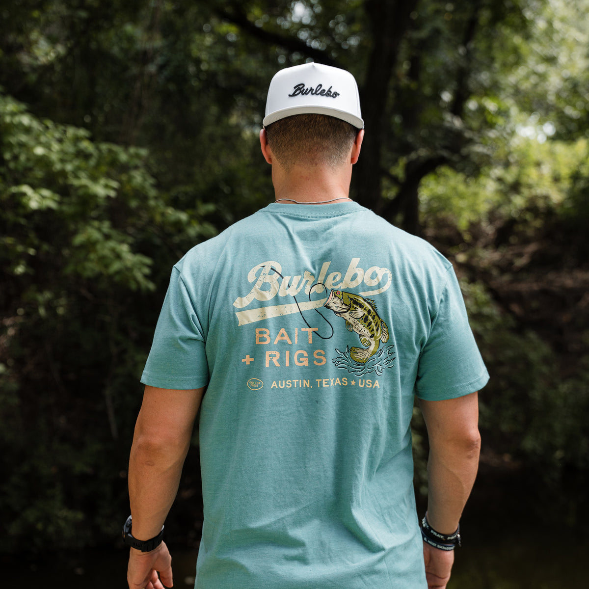 Man wearing a teal t-shirt with 'Burlebo' branding and a cap, standing in a forested area.