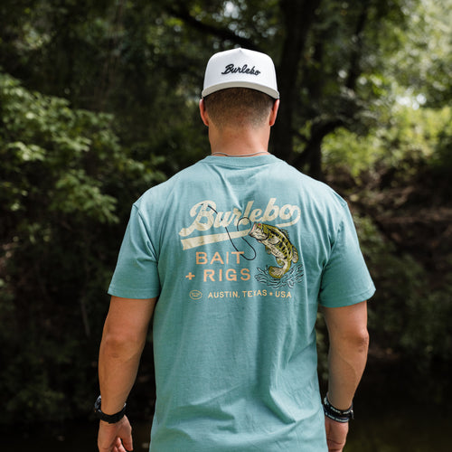 Man wearing a teal t-shirt with 'Burlebo' branding and a cap, standing in a forested area.