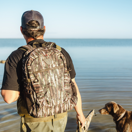 Man with camouflage backpack and cap standing on a beach with a dog, holding a duck.
