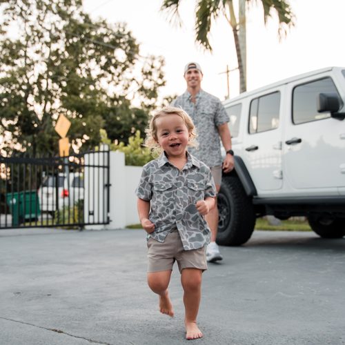 Child in camouflage shirt and shorts running in front of a white SUV with an adult behind.