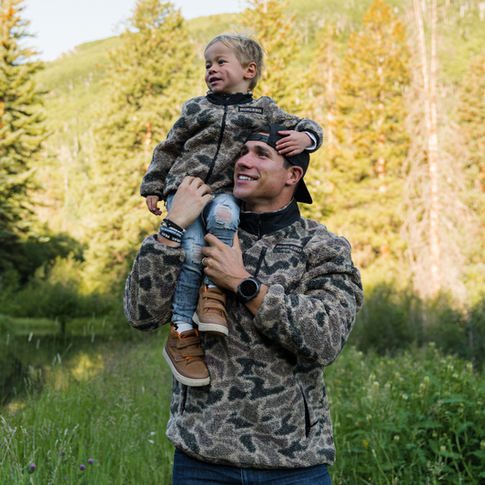 Man holding a child in a forested area with trees and grass in the background