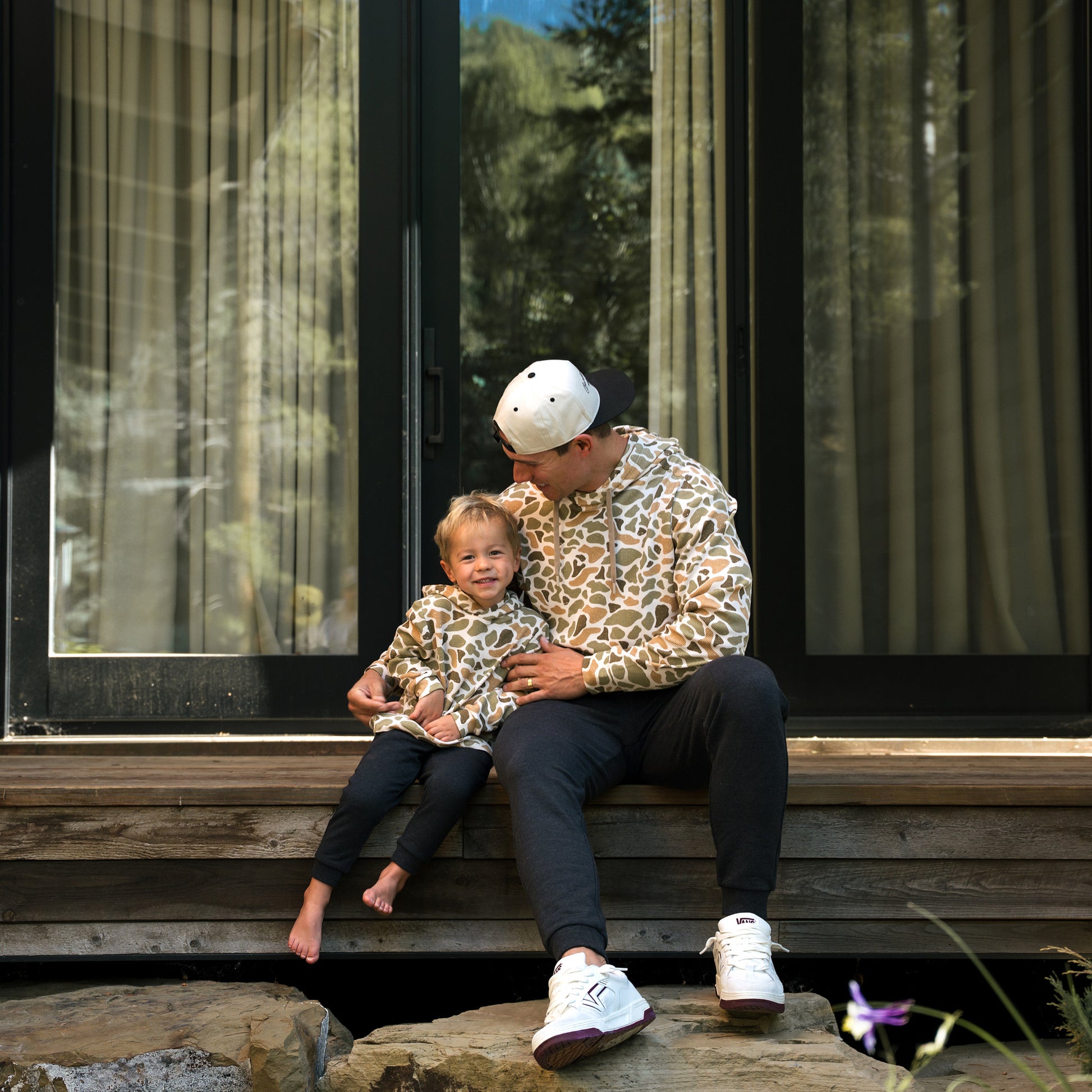 Man and child sitting on a wooden ledge outdoors with large windows in the background