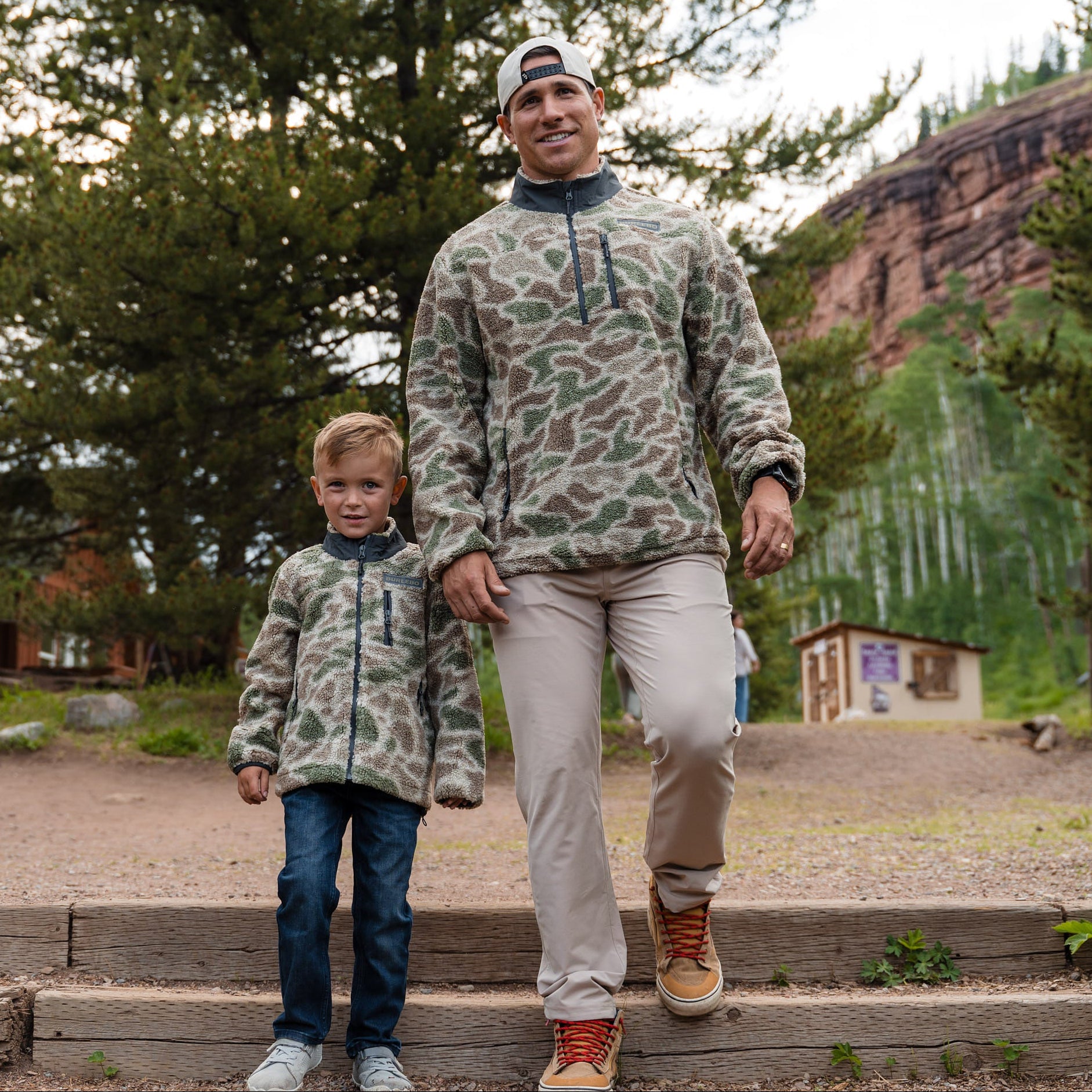 Man and child in matching camouflage outfits walking outdoors with trees and a building in the background.