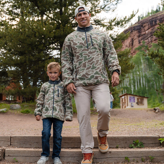 Man and child in matching camouflage outfits walking outdoors with trees and a building in the background.