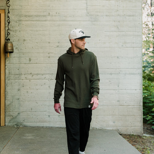 Man in a white cap walking past a concrete wall in the Mallard Green Thermal Hoodie