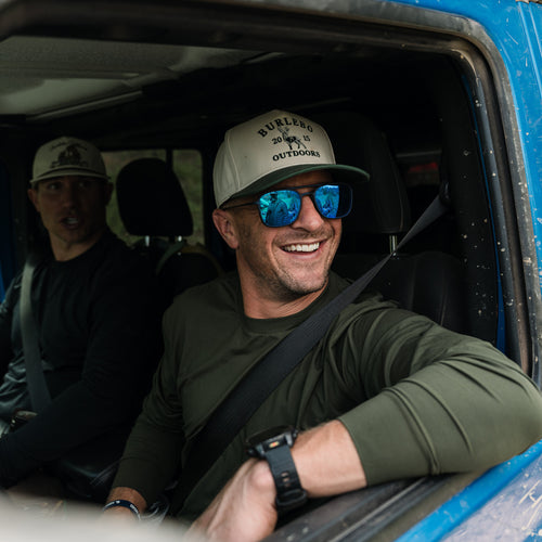 Smiling man in sunglasses and a cap wearing the Mallard Green Long Sleeve Go To Tee in a blue off-road vehicle