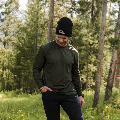 Smiling man in a black beanie wearing the Mallard Green Long Sleeve Go To Tee in a pine forest